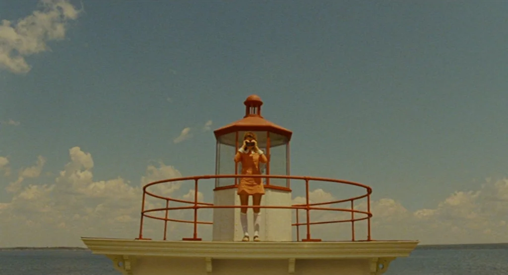 Moonrise Kingdom (2012) movie still: wide — A girl stands on the balcony of a lighthouse surrounded by a large body of water. She looks through…