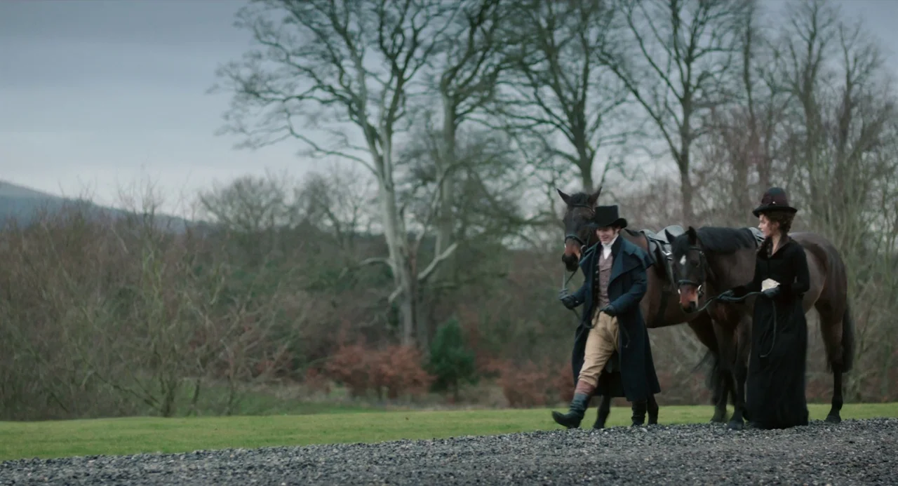 Love and Friendship (2016) movie still: fullbody — A man and a woman in 19th-century riding attire walk along a gravel path. They lead two horses…