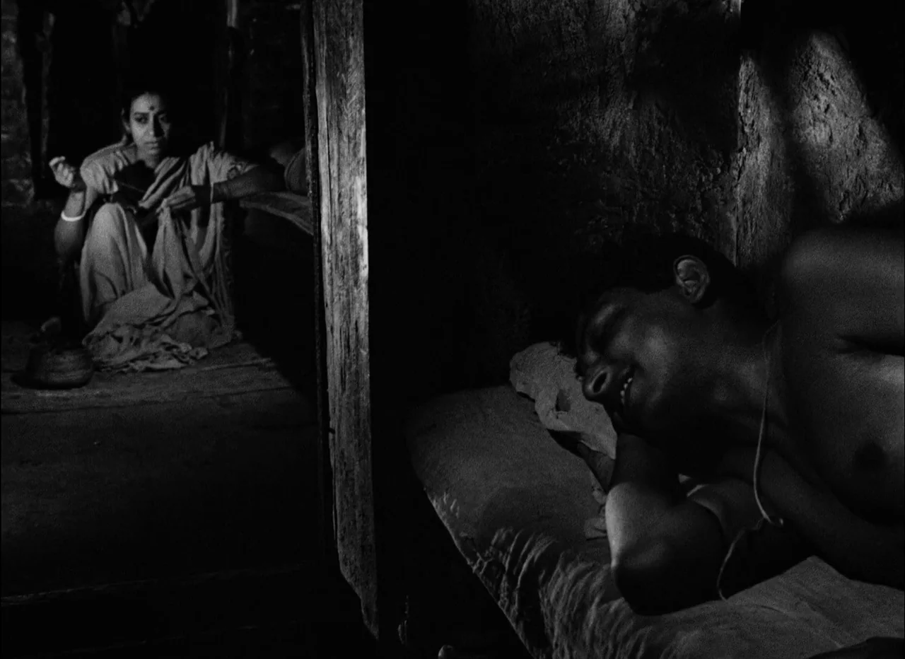 Pather Panchali (1955) movie still: medium — A dimly lit, divided mud interior features a man sleeping and a woman working. The structural division…