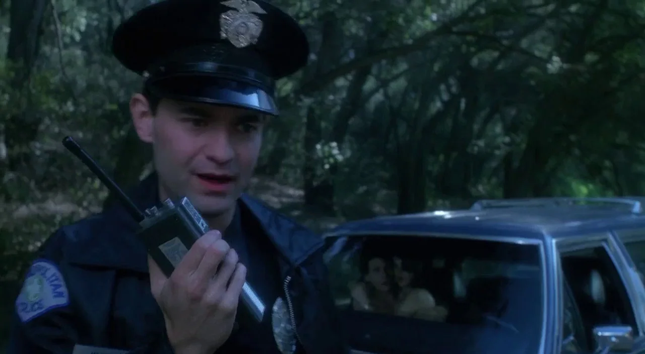 Heathers movie still: medium — A dark-uniformed officer stands on a tree-lined road near a parked station wagon. The man holds a black two-way radio…