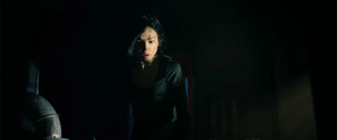 Ash (2025) movie still: medium — A dark industrial space features a curved metal pipe in the immediate foreground. A young woman stands in the…