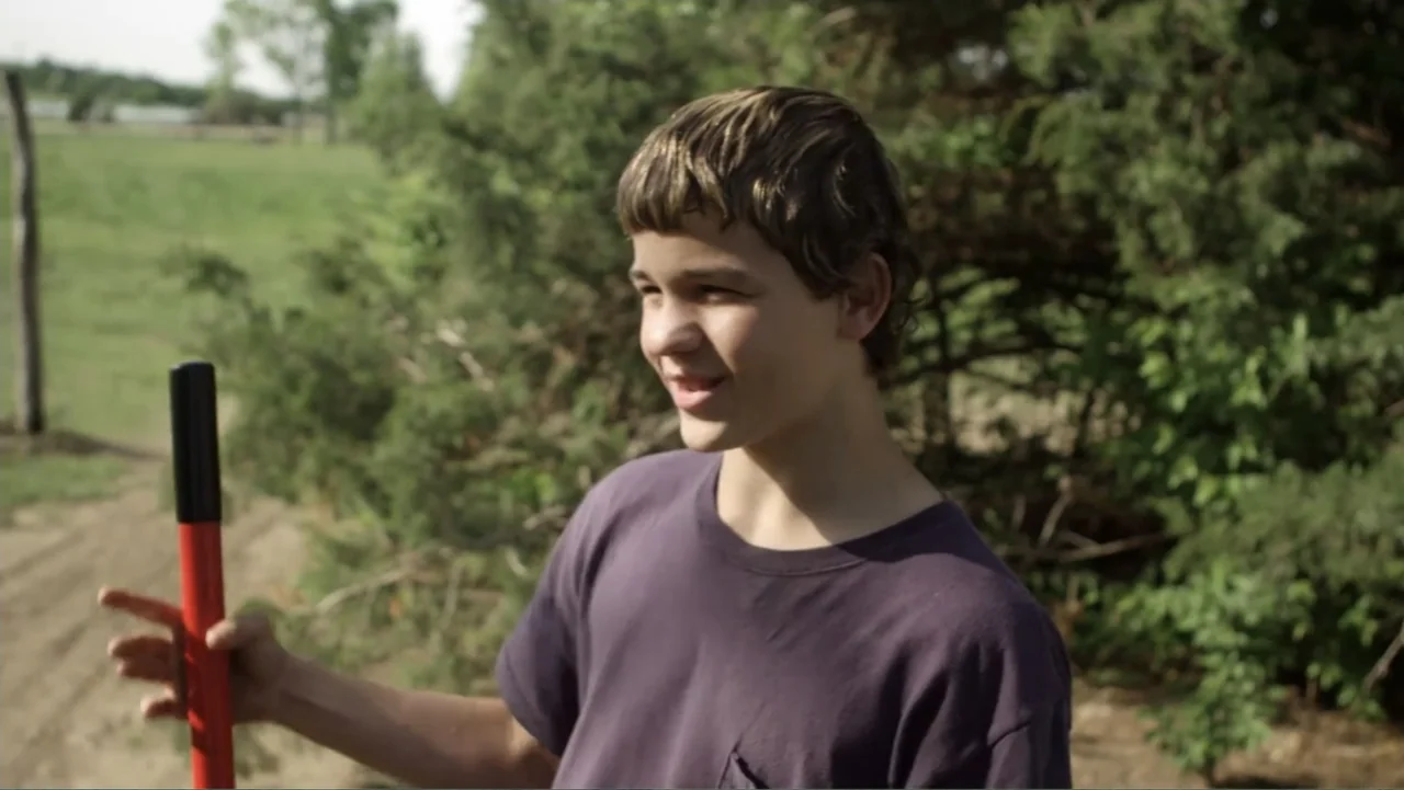 Rich Hill (2014) movie still: medium — A rural field with scattered evergreen trees and dry earth under a clear sky. A young boy in a purple t-shirt…