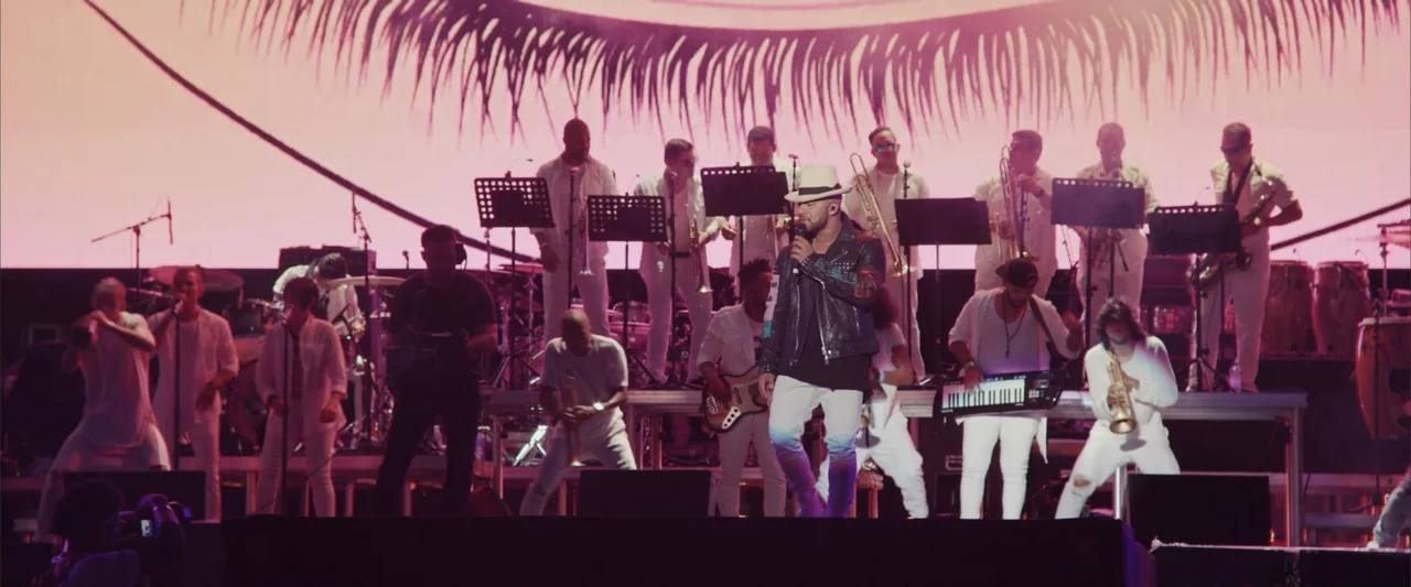 La Musiquita por Dentro (2019) movie still: fullbody — A crowded musical performance occurs on a well-lit stage with a giant graphic backdrop.…