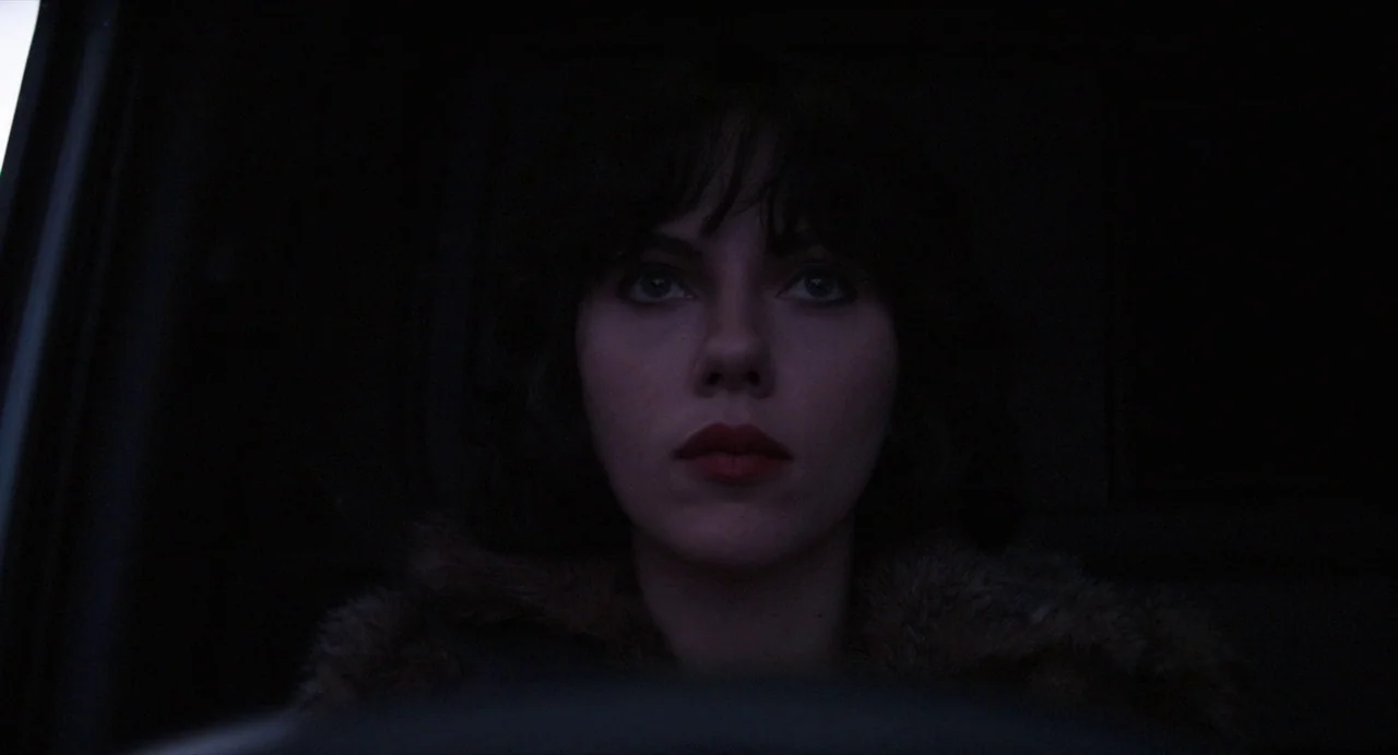 Under The Skin (2013) movie still: closeup — The interior of a vehicle is dark with only faint illumination on the driver. A woman sits behind the…