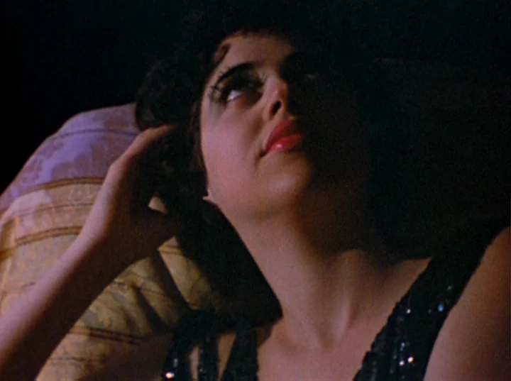 Puce Moment (1949) movie still: A woman reclines in the dark, her face partially illuminated by a soft, warm light that accentuates her heavy makeup…