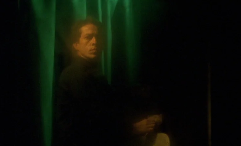 The Double Life of Véronique (1991) movie still: medium — A man stands in a dark space positioned behind a large, folded green curtain. He stares…