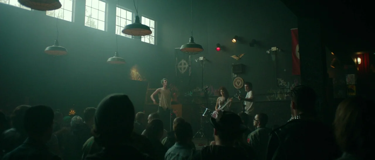 Green Room (2015) movie still: wide — A dark, smoky concert space features a small stage with a performing band. The walls are covered in graffiti…