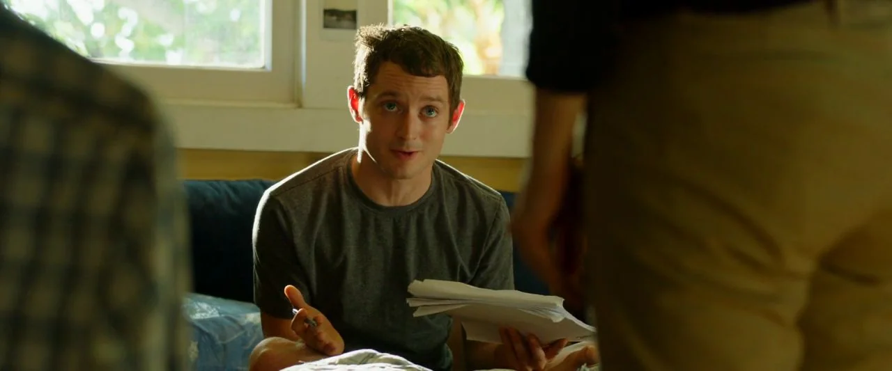 Cooties (2014) movie still: medium — An interior space features a man seated in front of a white window frame. The man holds a stack of white papers…