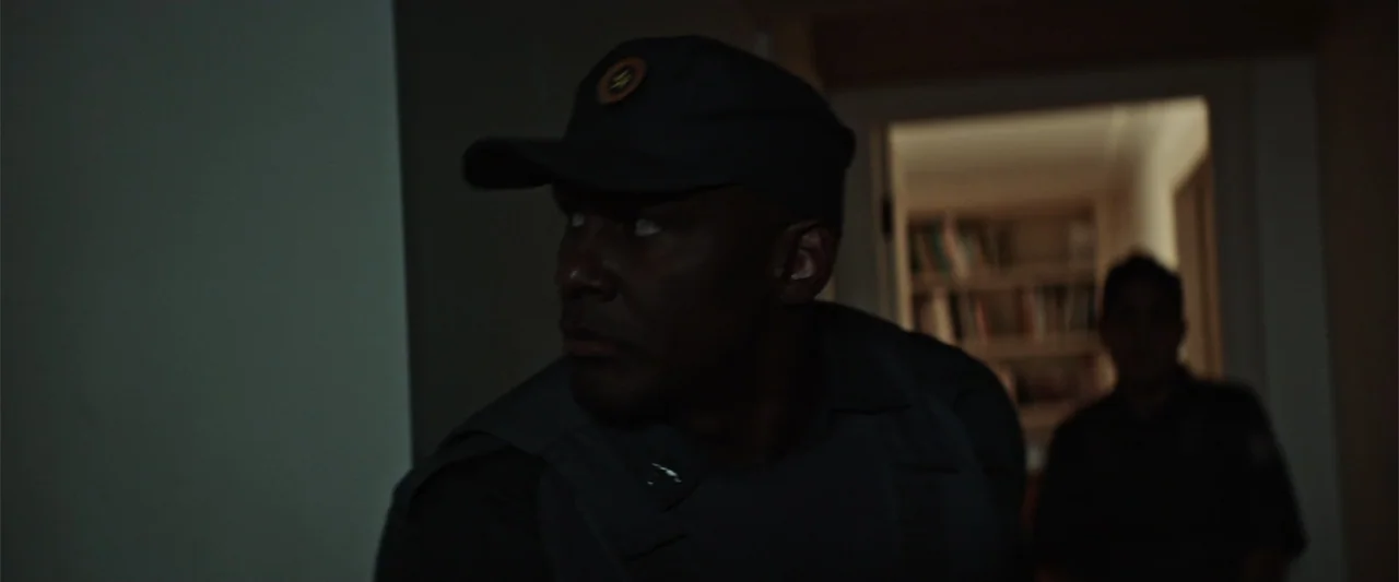 The Boy Who Killed My Parents (2021) movie still: medium — A dimly lit indoor hallway with a blurred bookshelf visible through a doorway. A man in a…