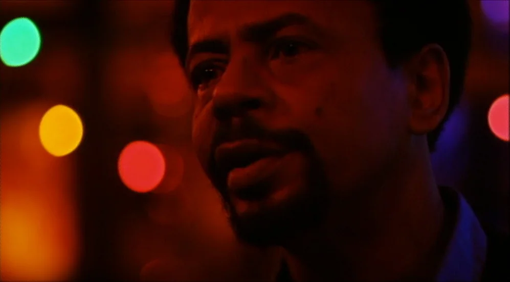 Clockers (1995) movie still: closeup — A dimly lit indoor space features blurred, vibrant bokeh lights in the background. A man is positioned in the…