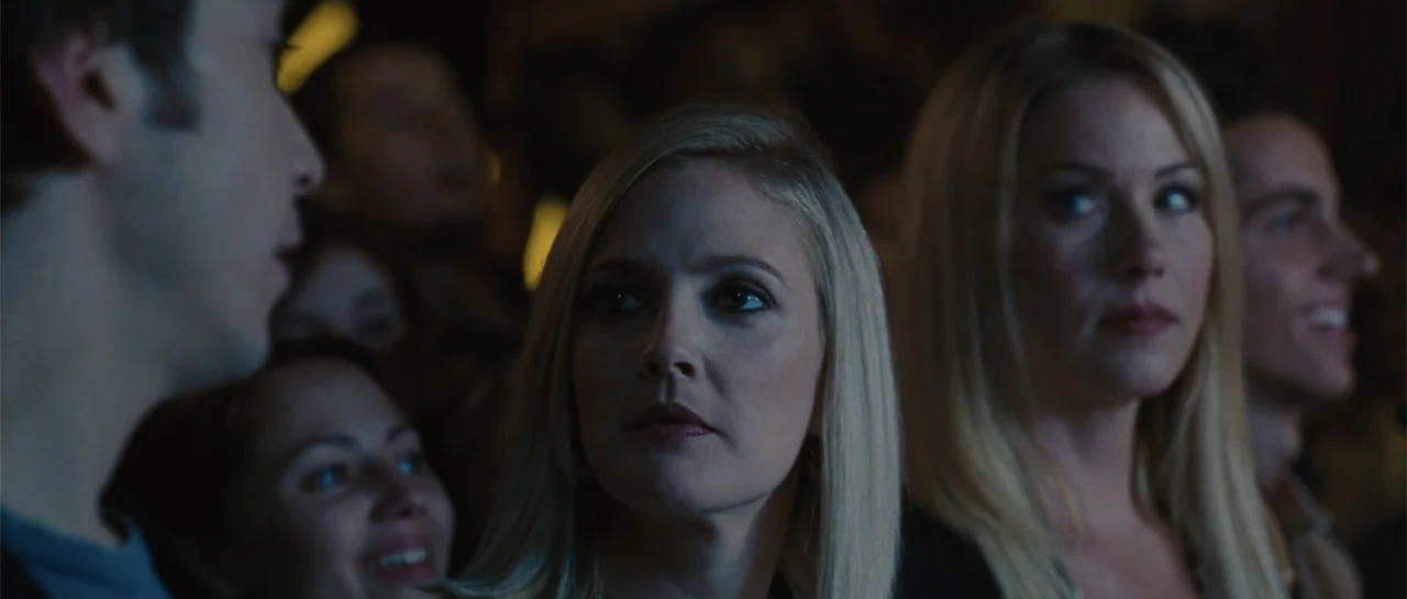 Going the Distance (2010) movie still: medium — A dark indoor auditorium with multiple people seated closely together facing a screen. Two blonde…