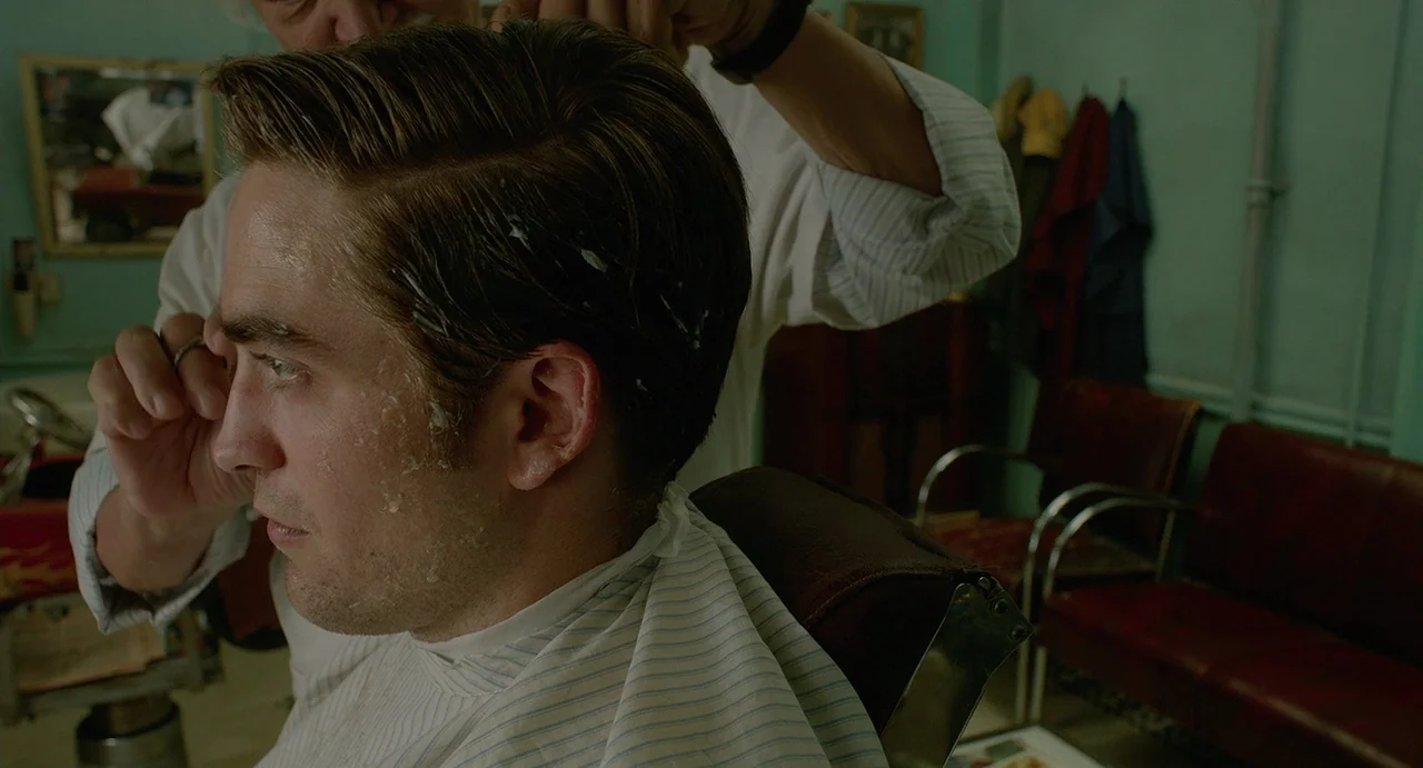 Cosmopolis (2012) movie still: closeup — A man sits in a vintage chair inside a small room with light green walls. A barber applies styling product…