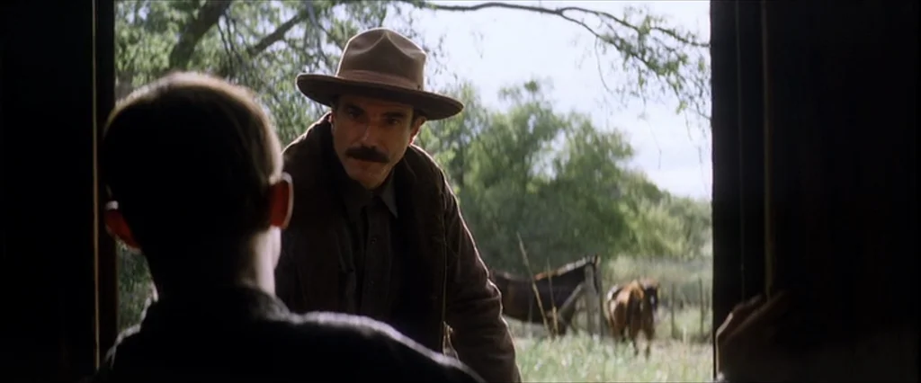 There Will Be Blood (2007) movie still: medium — A man stands in a wooden doorway overlooking a field with horses. The scene is lit by natural…