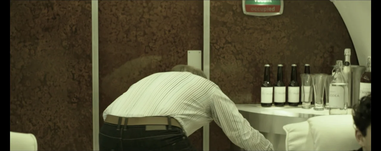 Turbulence (2025) movie still: medium — A narrow airplane galley with polished wood-paneled walls and a small service counter. A man in a striped…