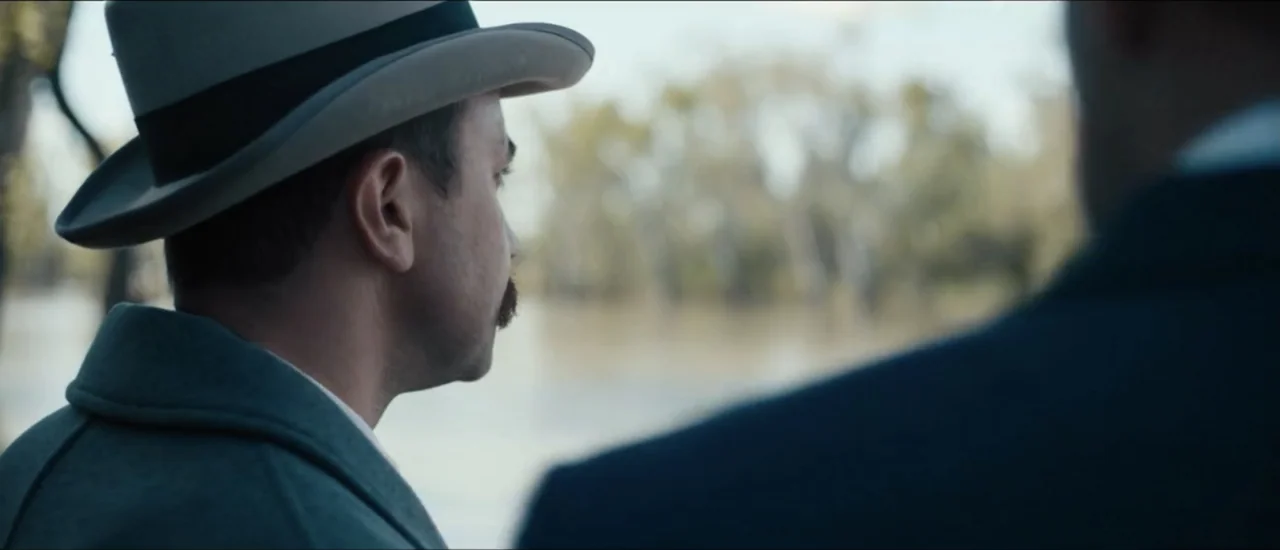 Fear Below (2025) movie still: medium — A man in a fedora stands by a muddy riverbank flanked by pale trees. He looks out across the water while a…