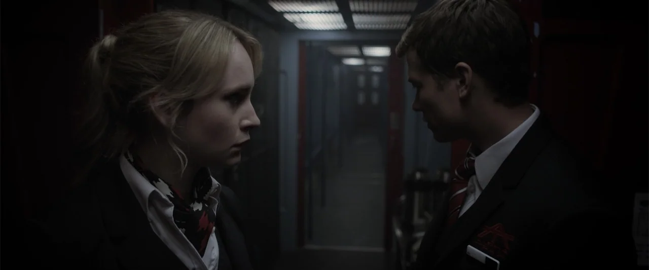Howl (2015) movie still: medium — A narrow service corridor features dark walls and overhead lights set in an airplane. A man and a woman stand face…