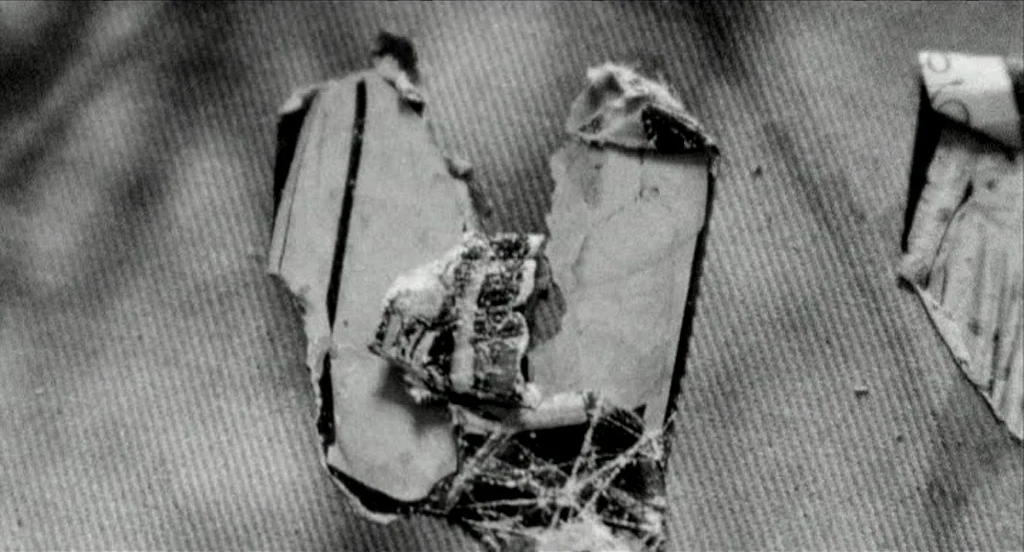 Container (2006) movie still: closeup — A torn, crumpled piece of paper rests on a coarse, fibrous surface. The paper fragment shows remnants of…