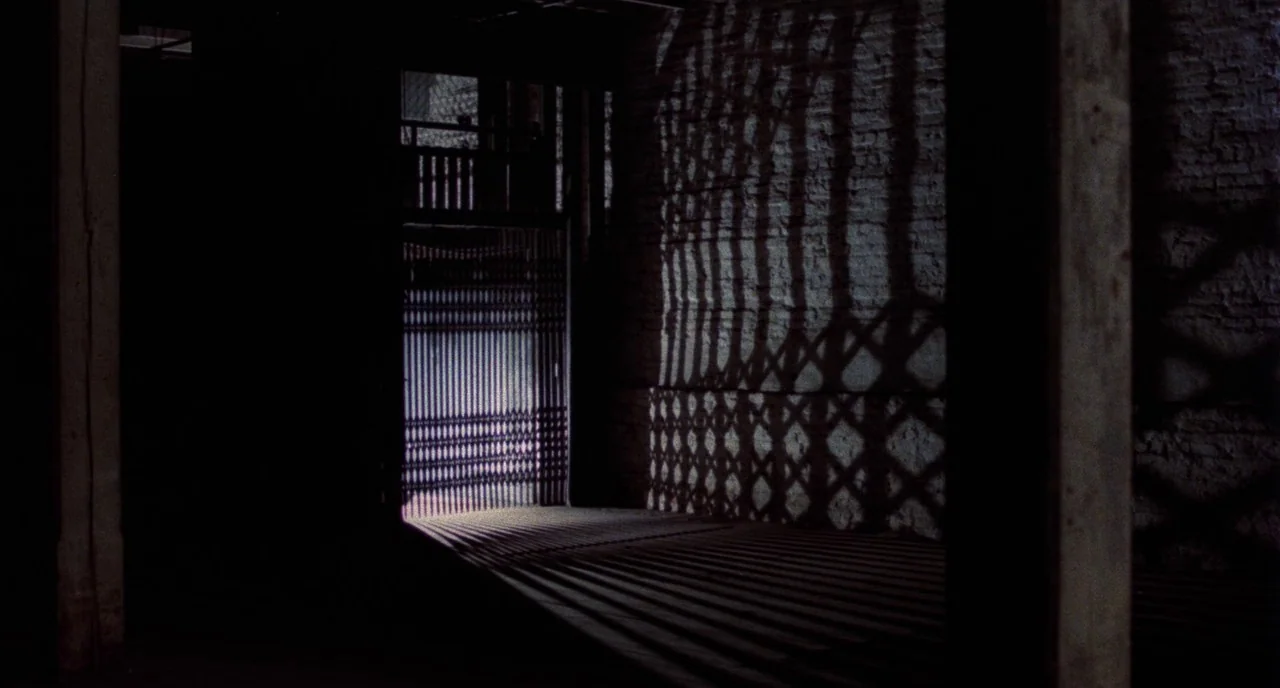 Angel Heart (1987) movie still: establishing — A dark industrial space features an iron gate illuminated from behind. Long geometric shadows stretch…