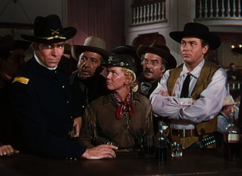 Calamity Jane (1953) movie still: medium — A group stands together inside a 19th-century frontier bar with wooden decor. They are gathered around a…