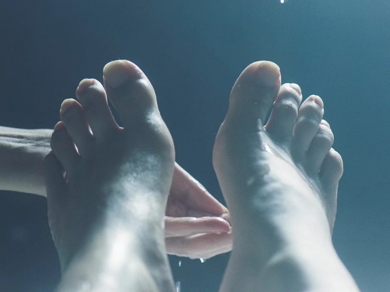 Joyland (2022) movie still: closeup — A dark, enclosed space containing a basin of water is illuminated by cool light. A pair of bare feet are…
