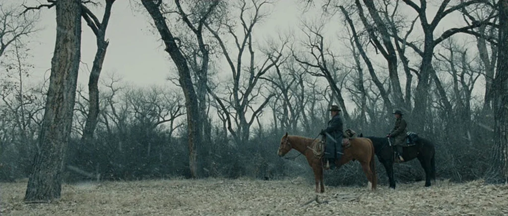 True Grit (2010) movie still: wide — A dense grove of leafless deciduous trees stands in a sparse, snowy landscape. Two men ride horses across the…
