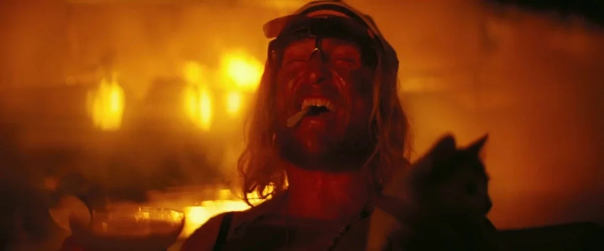 The Beach Bum (2019) movie still: closeup — A man stands amidst an intensely bright orange firelit environment with heavy smoke. The man has long…