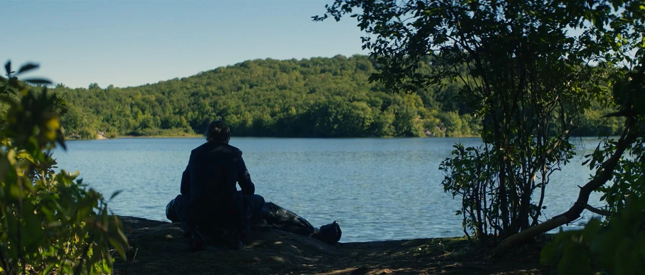You Were Never Really Here (2017) movie still: wide — A man sits on a rocky shoreline facing a calm, expansive lake. Densely wooded green hills line…