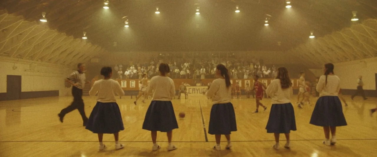 The Vast of Night (2019) movie still: wide — A high school gymnasium with wooden floors, bleachers, and rows of suspended stadium lights. A group of…