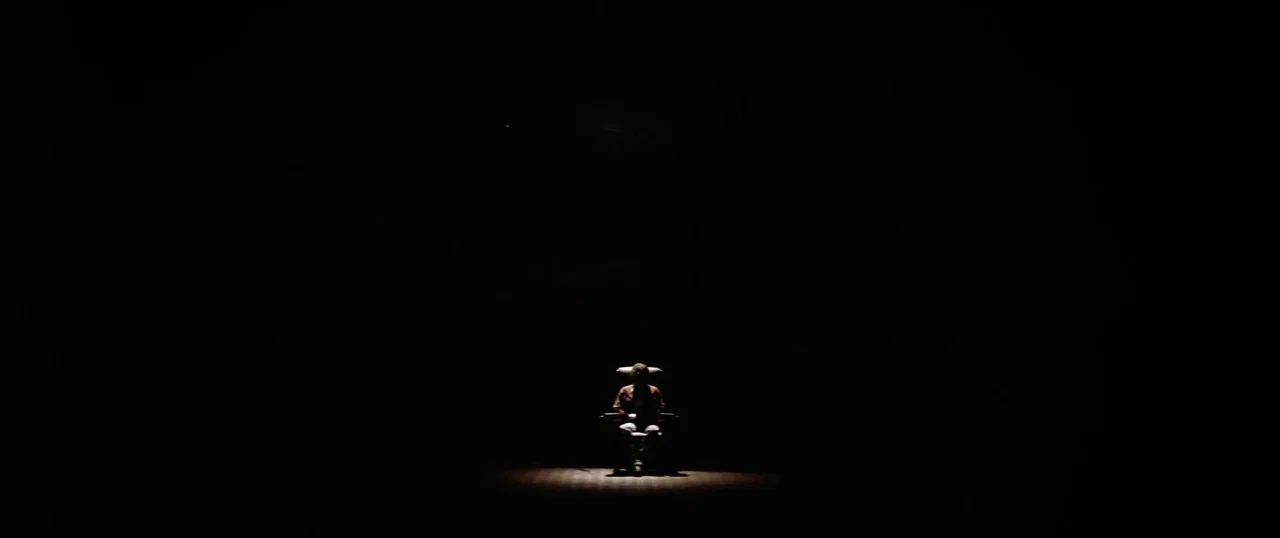 The Parallax View (1974) movie still: wide — A dark, empty stage is defined only by a sharp circle of overhead light. A lone man sits on a chair in…