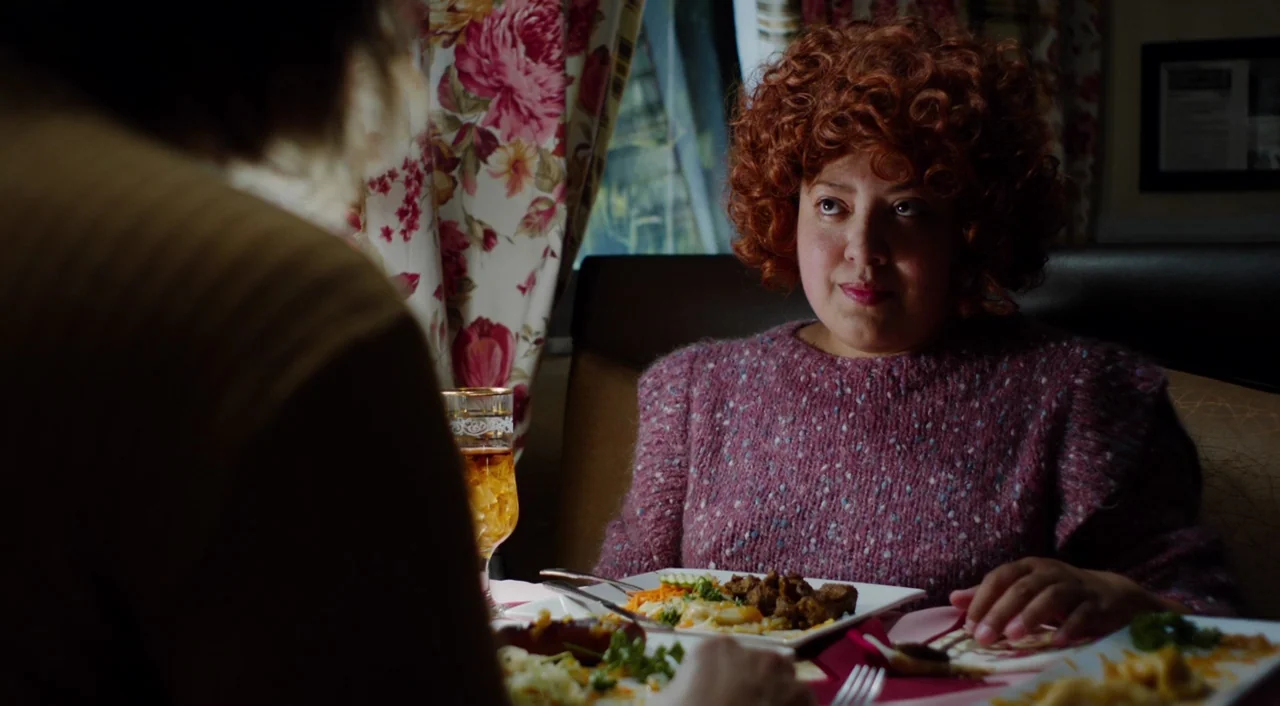 The Greasy Strangler (2016) movie still: medium — A dark booth with floral curtains behind a woman in a thick sweater. She sits across from another…
