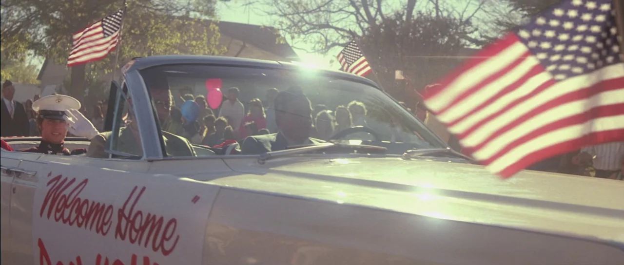 Born On The Fourth of July (1989) movie still: medium — A convertible car decorated for a parade moves through a residential street. A soldier in…