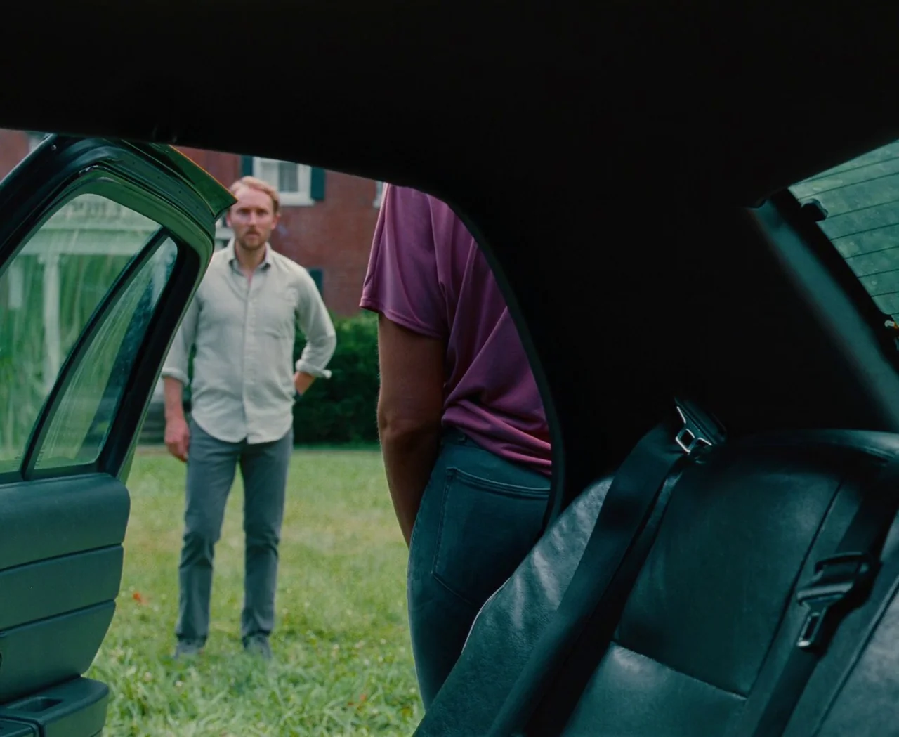 Monica (2022) movie still: medium — A green lawn sits before a brick house on a bright day. A man stands in the grass as a person exits the back of…