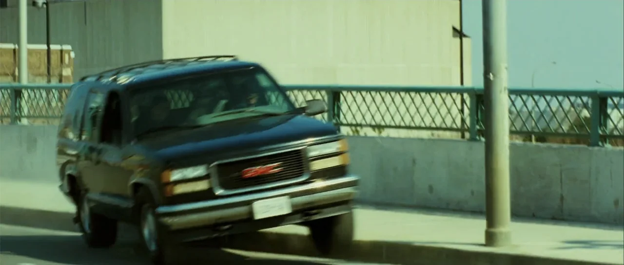 Shoot 'Em Up (2007) movie still: medium — A dark GMC SUV drives along a concrete bridge with a green metal railing. The vehicle moves from the left…