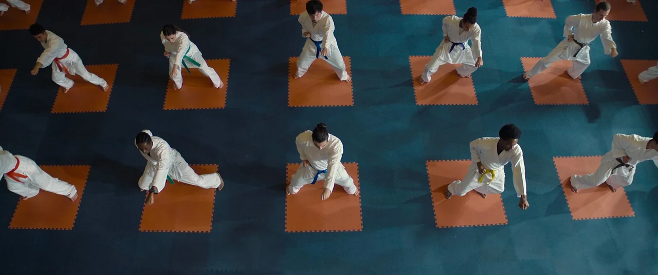 Polite Society (2023) movie still: wide — A group of martial arts students train in an interior studio with blue flooring. Each student stands on an…