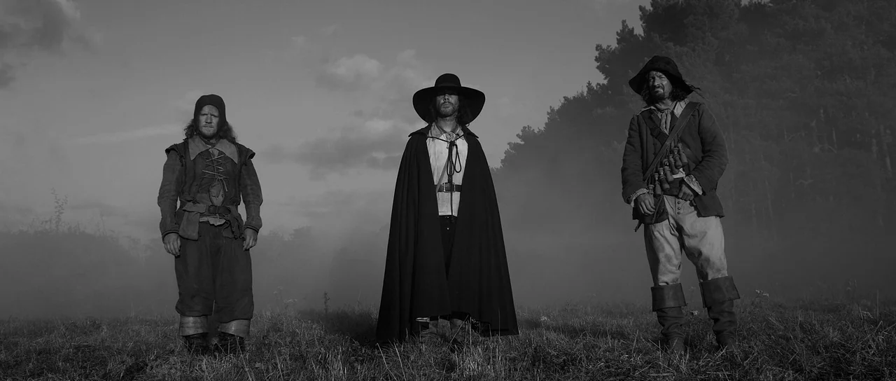 A Field In England (2013) movie still: fullbody — Three men dressed in seventeenth century attire stand in an open field of tall grass. Fog hangs…