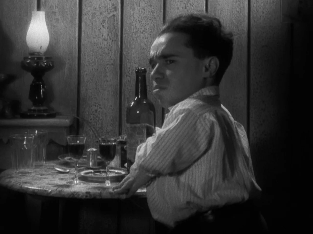 Freaks (1932) movie still: medium — A wood-paneled tavern corner features a small round table with glassware and a bottle. A man sits by the table…