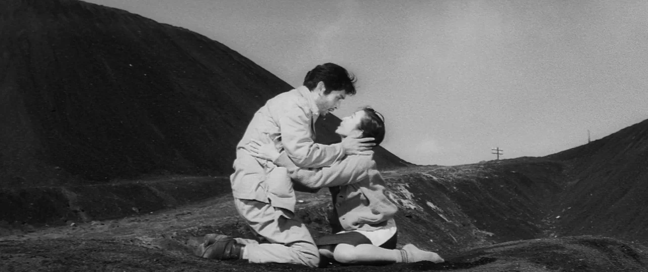 The Human Condition I: No Greater Love (1959) movie still: fullbody — A man and woman kneel facing each other on a dark, barren industrial hill. The…
