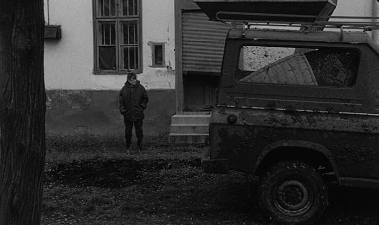 Sátántangó (1994) movie still: fullbody — A rural house exterior features a weathered building with a rolled-down shutter and grass. A woman in a…
