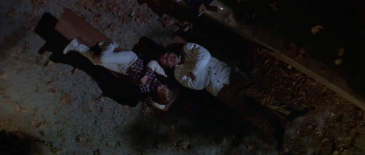 Invaders From Mars (1986) movie still: wide — Two individuals lie on a wooden bench at night in a patch of park. Fallen leaves are scattered across…