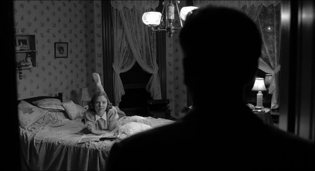 The Man Who Wasn’t There (2001) movie still: medium — A dark bedroom with floral wallpaper and lace curtains features a woman in bed. A man in the…