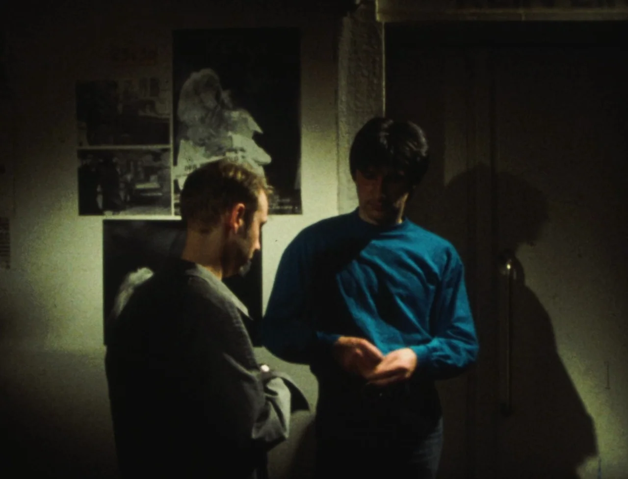 Nekromantik (1988) movie still: medium — Two men stand in a dim interior area against a wall adorned with posters. The figures are illuminated by an…