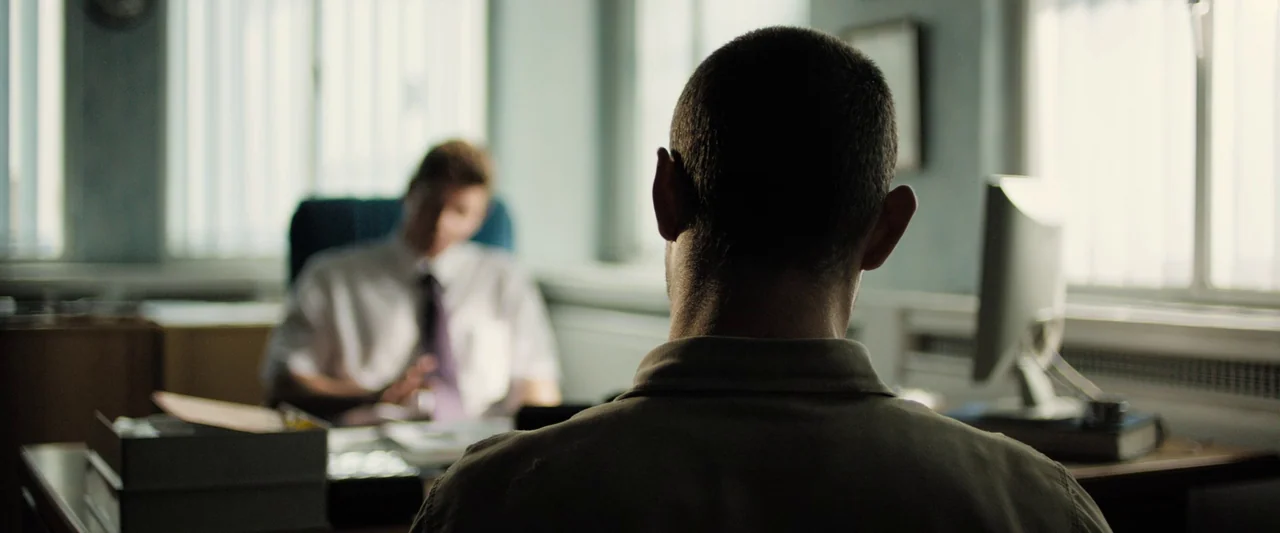Black Sea (2014) movie still: medium — A utilitarian office features white walls, window blinds, and a desktop computer monitor. A man in the…
