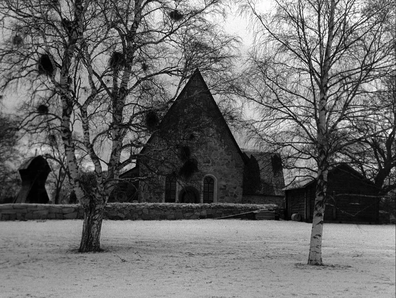 Winter Light (1963) movie still: establishing — A stone church with a steeply pitched roof stands behind a low stone wall. Leafless birch trees…