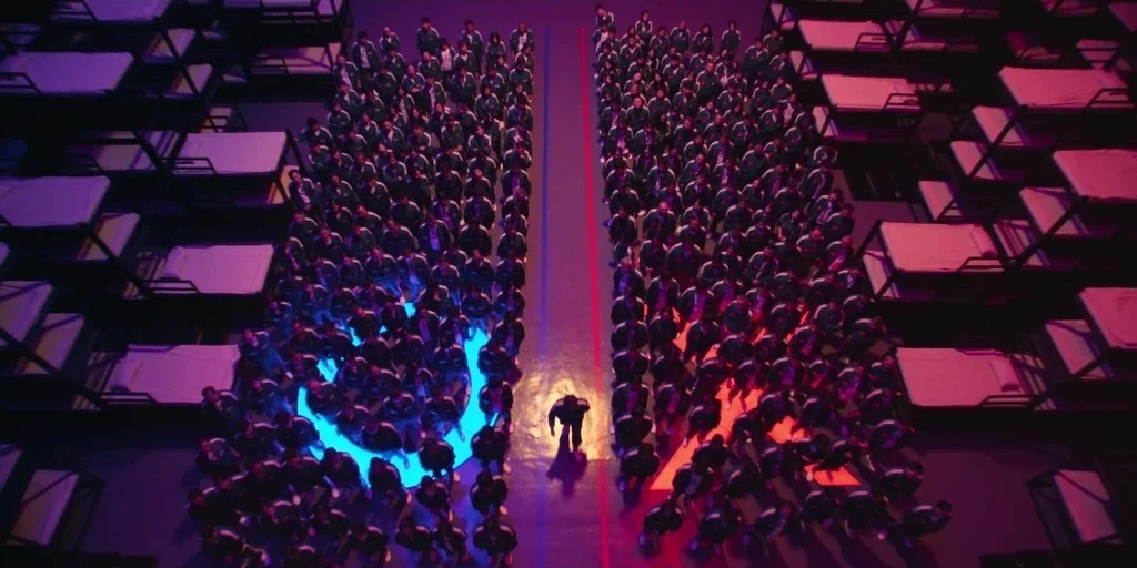 Squid Game (2024) movie still: wide — A vast, sterile industrial dormitory filled with stacks of empty metal bunk beds. Large crowds of people stand…