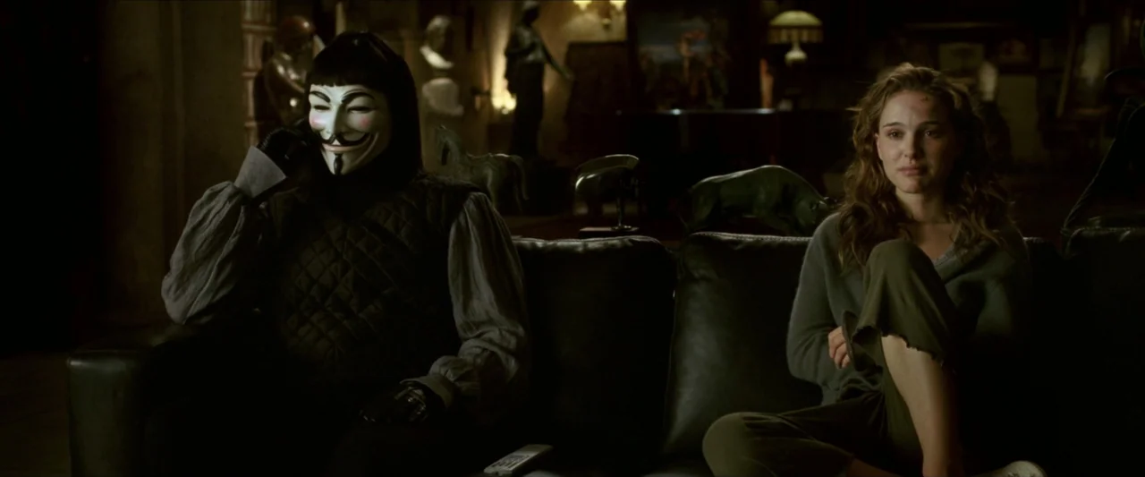 V for Vendetta (2006) movie still: medium — A dimly lit room contains a dark leather sofa and various decorative statues. A masked figure sits next…