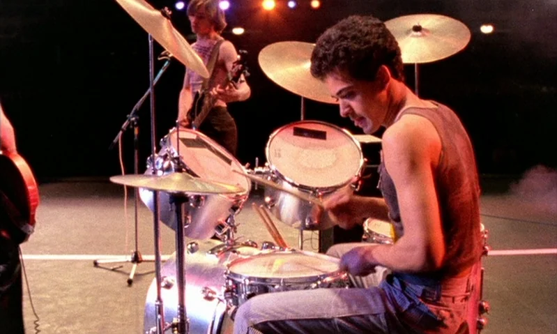 Jubilee (1978) movie still: medium — A dark concert venue with a drum kit prominently displayed in the center. A young man sits behind the drums…