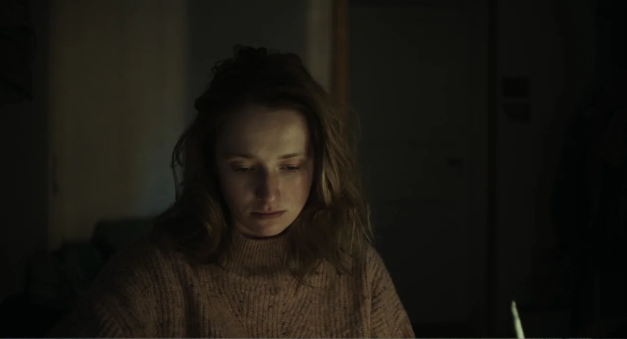Ninjababy (2021) movie still: closeup — A dark indoor space with minimal visible detail surrounding the subject. A woman sits in shadow while soft…