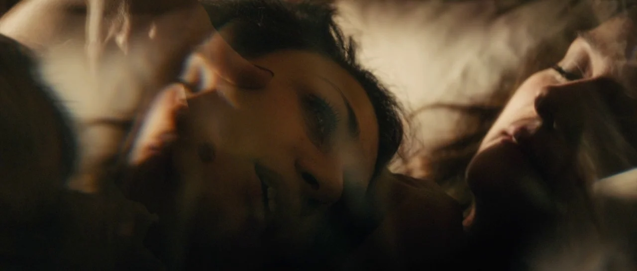 The Duke of Burgundy (2014) movie still: closeup — A dimly lit bed with white sheets and soft fabric pillows. Two young women lie side by side with…