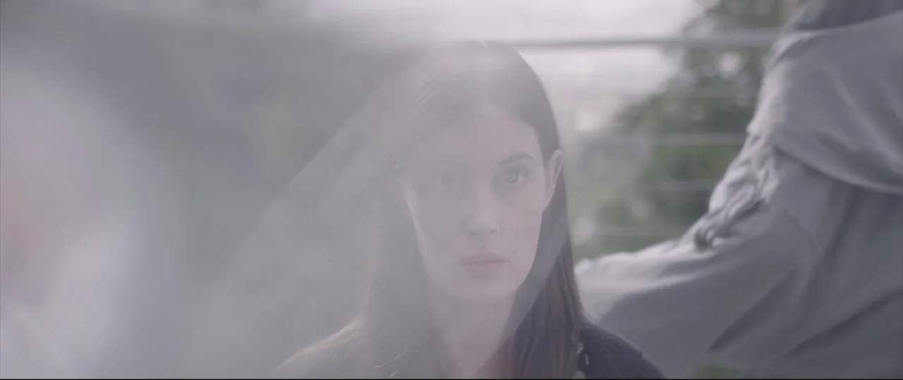Sun Choke (2015) movie still: closeup — A woman stands on an outdoor balcony with blurred greenery and railings in the background. She faces forward…