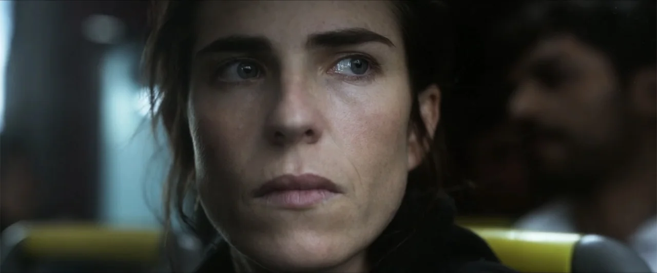 Dive (2013) movie still: closeup — Interior of a dark public transit vehicle with a blurred passenger in the background. A woman in a dark jacket…